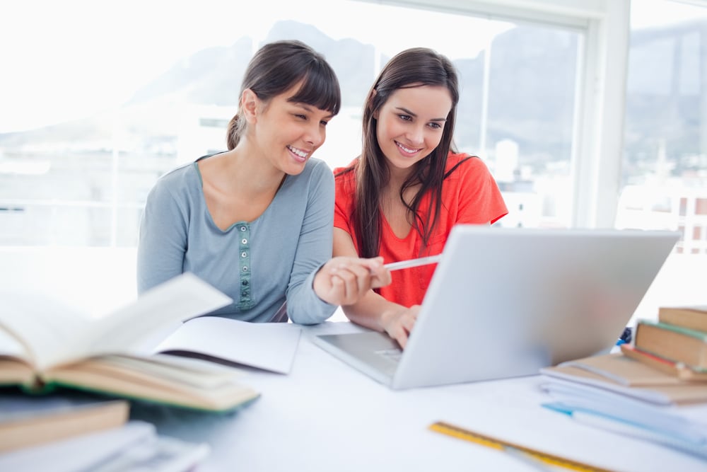 The girls are both sitting beside each other and smiling as they use the laptop as one girl points to something on screen